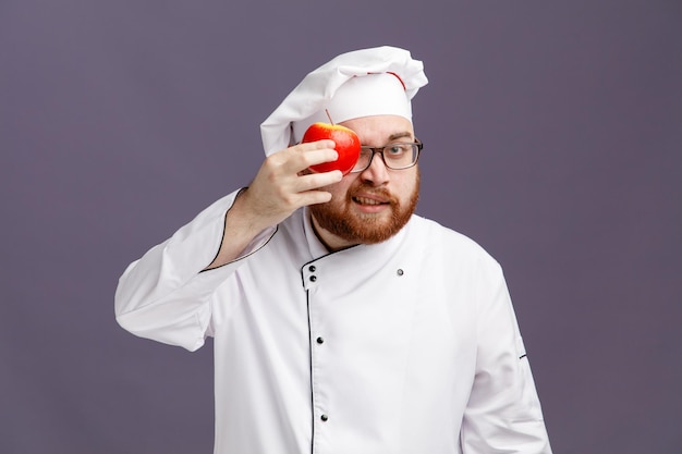 Foto fiducioso giovane chef con gli occhiali uniforme e cappuccio che tiene la mela davanti all'occhio guardando la fotocamera isolata su sfondo viola