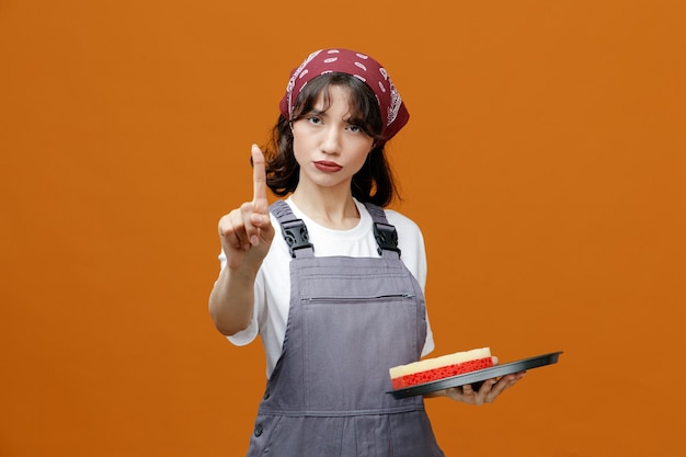 Confident young female cleaner wearing uniform and bandana holding tray with sponge in it looking at camera showing hold on gesture isolated on orange background