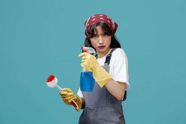 Fiduciosa giovane donna addetta alle pulizie che indossa bandana uniforme e guanti di gomma che tengono spazzola e detergente guardando la fotocamera isolata su sfondo blu