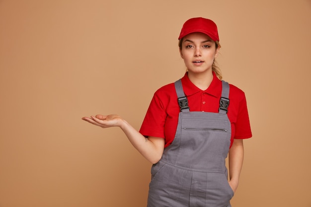 Confident young female construction worker wearing cap and uniform keeping hand in pocket showing empty hand 