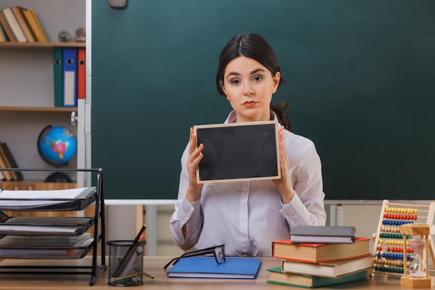 confident young female teacher holding mini blackboard sitting at desk with school tools in classroom