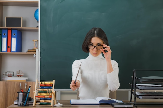 confident young female teacher wearing glasses points at camera with pointer sitting at desk with school tools on in classroom