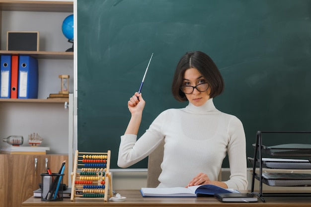 confident young female teacher wearing glasses points with pointer at blackboard sitting at desk with school tools on in classroom