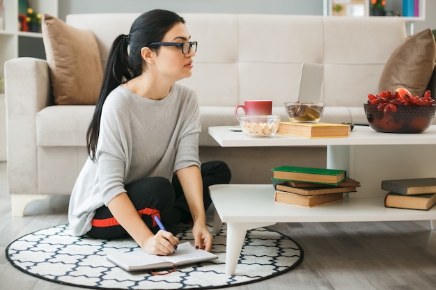 Confident young girl wearing glasses used laptop writes on notebook sitting on floor behind coffee table in living room