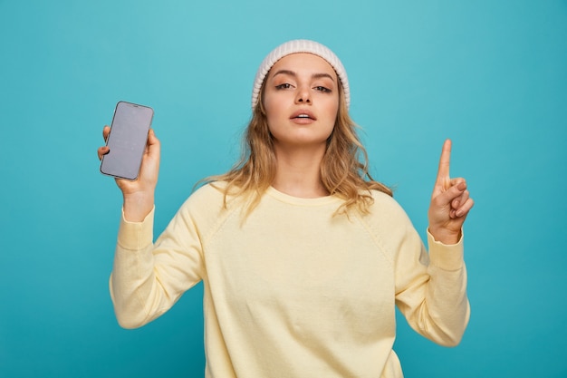 Confident young girl wearing winter hat holding mobile phone pointing up 