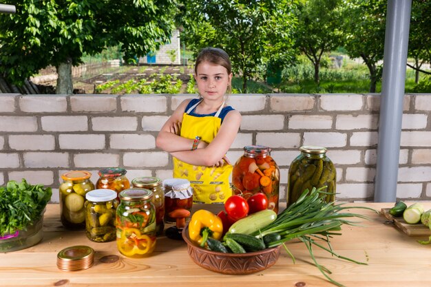 Confident young girl with her assorted preserved fresh vegetables displayed in glass jars on the table in front of her