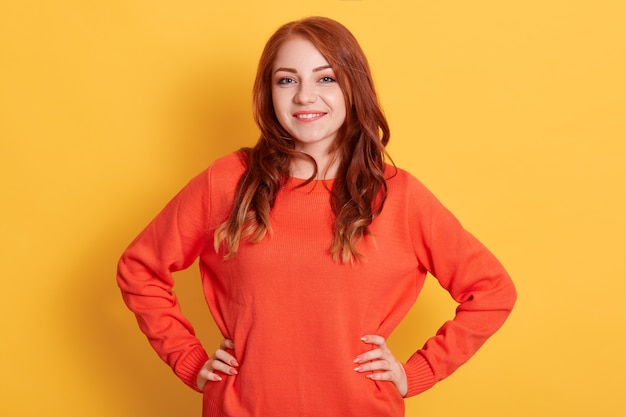 Confident young lady with red hair standing with hands on hips and looking 