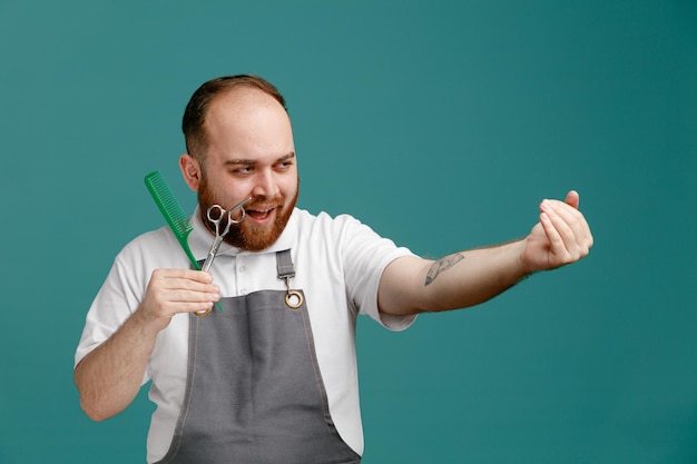 Confident young male barber wearing white shirt and barber apron showing comb and scissors looking at side showing come here gesture isolated on blue background