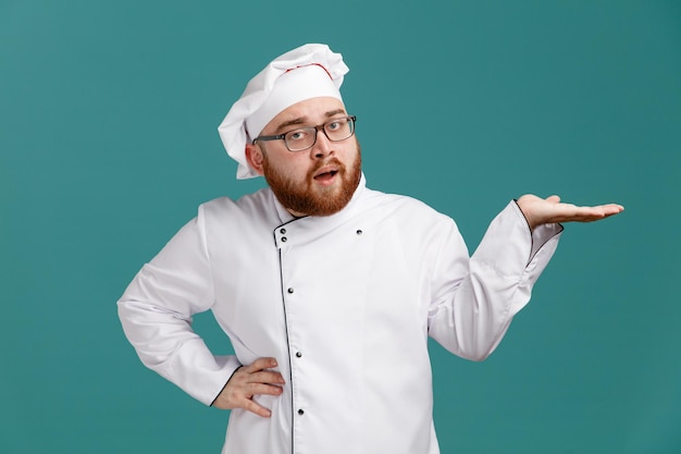 Уверенный молодой шеф-повар в очках и кепке смотрит в камеру, показывая пустую руку, держа другую руку на талии изолированной на синем фоне