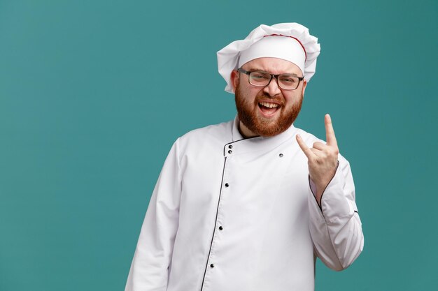 Confident young male chef wearing glasses uniform and cap looking at camera showing rock sign isolated on blue background