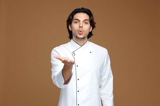 confident young male chef wearing uniform looking at camera showing kiss gesture and empty hand isolated on brown background