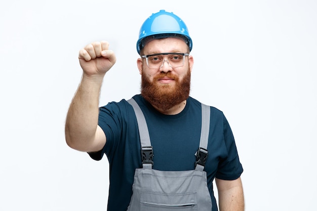 Confident young male construction worker wearing safety helmet uniform and safety glasses looking at camera showing knocking gesture isolated on white background