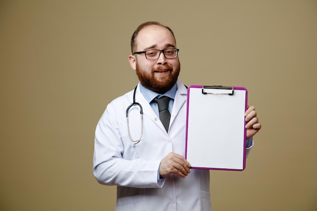 Confident young male doctor wearing glasses lab coat and stethoscope around his neck looking at camera showing clipboard isolated on olive green background