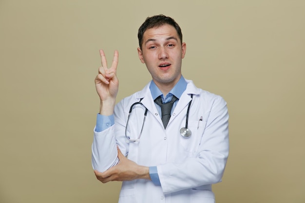 Confident young male doctor wearing medical robe and stethoscope around neck grabbing his arm looking at camera showing peace sign isolated on olive green background