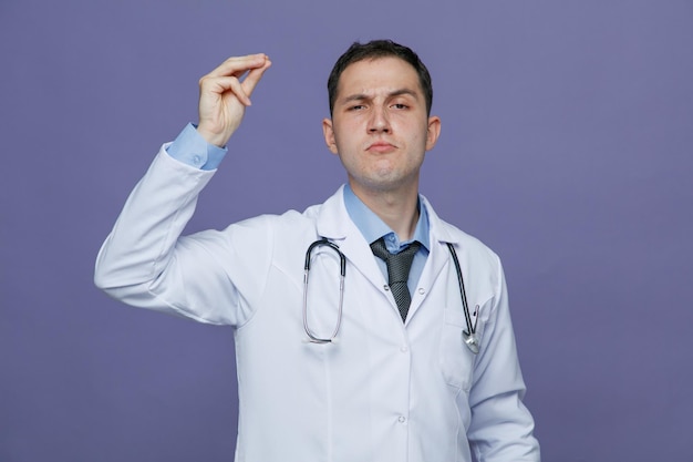 Confident young male doctor wearing medical robe and stethoscope around neck looking at camera making money gesture isolated on purple background