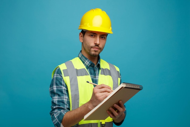 confident young male engineer wearing safety helmet and uniform holding pencil and note pad looking at camera isolated on blue background