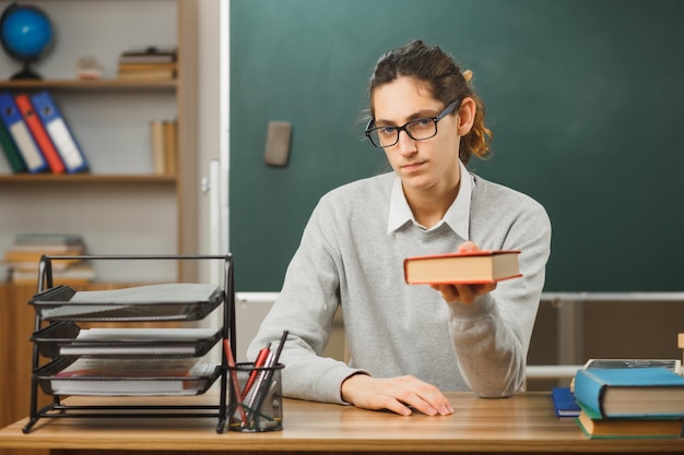 confident young male teacher holding out book at camera sitting at desk with school tools on in classroom