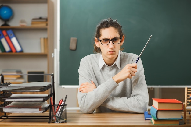 confident young male teacher wearing glasses holding pointer sitting at desk with school tools on in classroom