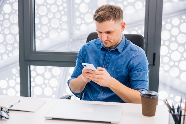 Confident young man in elegant casual clothes holds smartphone at his workplace in office