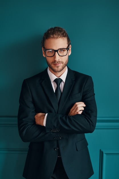 Photo confident young man in elegant suit looking at camera and keeping arms crossed while standing green wall