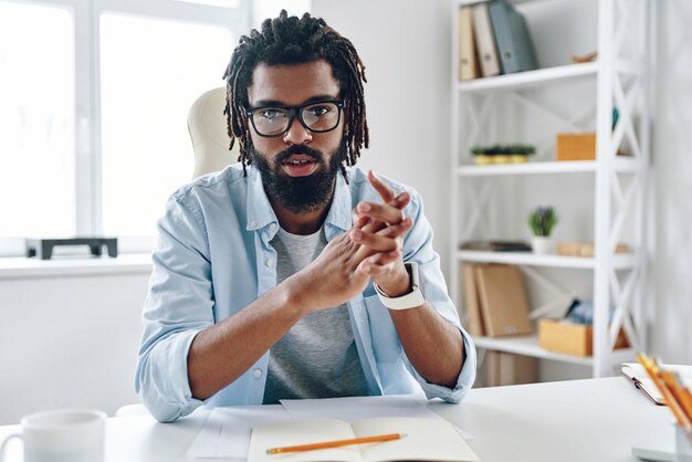 Confident young man in eyewear telling something and looking at camera while working indoors