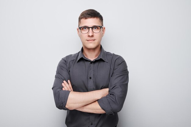 Confident young man in glasses and a gray shirt arms crossed standing against a plain background