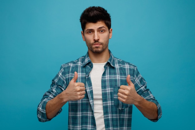 Confident young man looking at camera showing thumbs up isolated on blue background