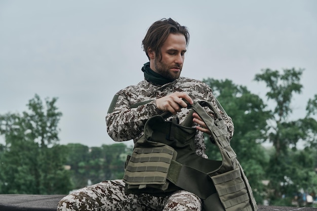 Foto giovane fiducioso in uniforme militare che si toglie l'attrezzatura mentre è seduto all'aperto