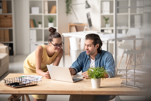 Photo confident young man presenting part of new project to colleague while both looking at laptop display at working meeting in office