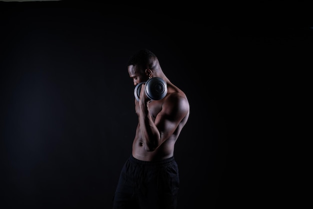 Confident young man shirtless portrait training with dumbbells against black background