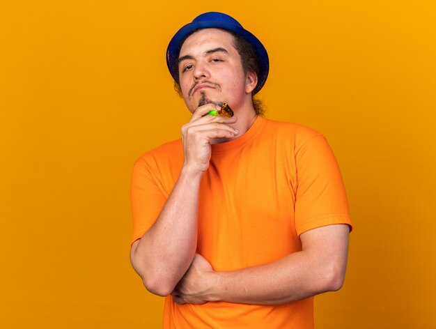 Confident young man wearing party hat holding party whistle holding hand on chin 