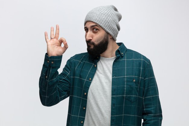 Confident young man wearing winter hat looking at camera showing ok sign isolated on white background