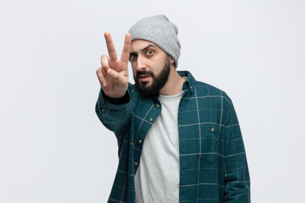 Confident young man wearing winter hat looking at camera stretching hand out towards camera showing peace sign isolated on white background