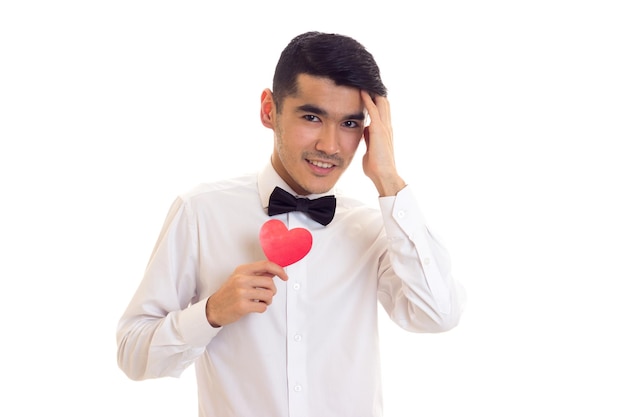 Confident young man with black hair in white T-shirt with black bow-tie holding a red paper heart on white background in studio