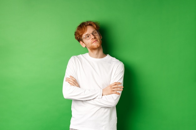 Confident young man with red hair, wearing glasses, looking smug at right side copy space, cross arms on chest self-assured, standing over green background.