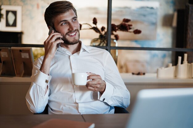 Confident young man working on laptop and talking on the mobile phone while sitting at his working place in office