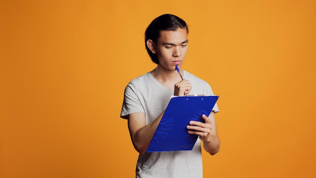 自信に満ちた若い男性が、ファイルにメモを書き、メモを取り、クリップボードと紙を使って情報を入手します。スタジオ、男性モデルで書類に署名するためにペンを持つ屈託のない現代人。
