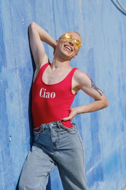 Foto fiducioso giovane persona non binaria che sorride mentre posa su una parete blu all'aperto in una giornata di sole. concetto di genere non binario.