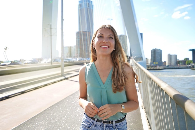 ロッテルダムオランダの日没時にエラスムス橋を歩く自信を持って若い笑顔の女性