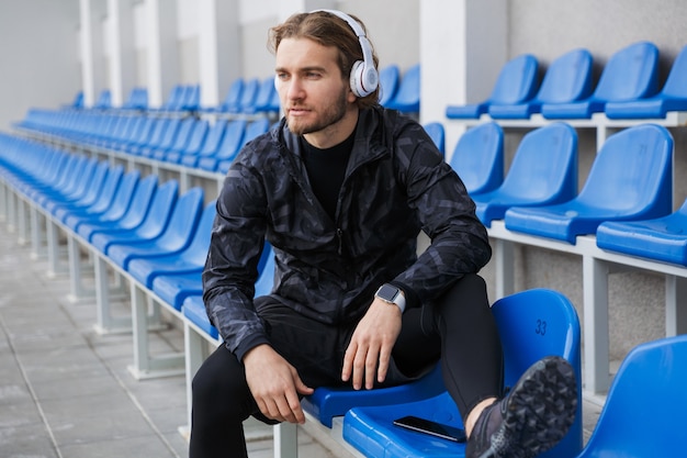 Confident young sportsman sitting on tribune seats at the stadium, listening to music with wireless headphones