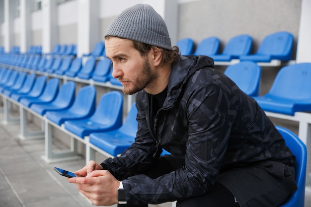 Confident young sportsman sitting on tribune seats at the stadium, using mobile phone