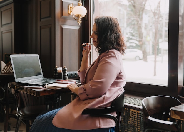 Confident young woman in a business suit works on a laptop while sitting by the window in a cafebusinesswoman