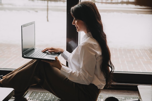 비즈니스 정장을 입은 자신감 있는 젊은 여성이 카페 사업가의 창가에 앉아 노트북 작업을 합니다