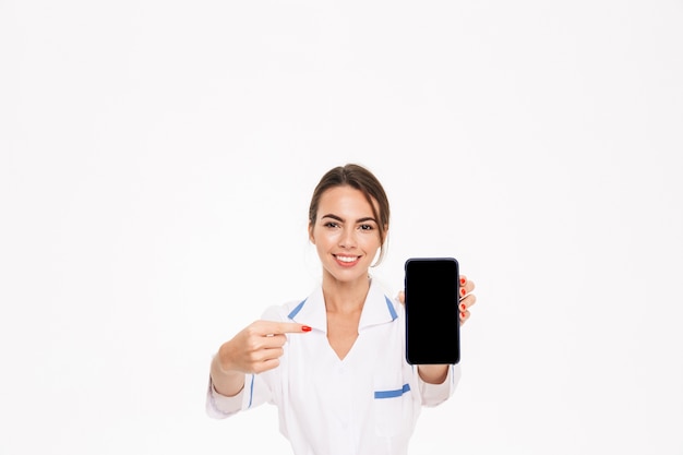 Confident young woman doctor wearing uniform standing isolated over white wall, showing blank screen mobile phone