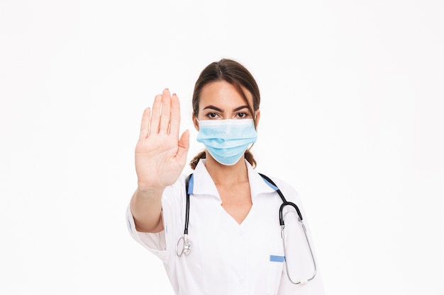 Confident young woman doctor wearing uniform standing isolated over white wall, showing stop gesture