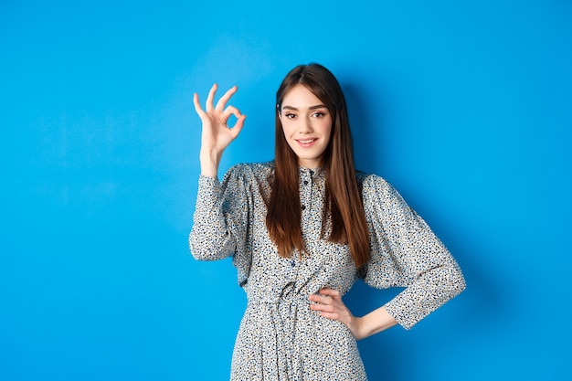 Confident young woman in dress showing okay sign and smiling, no problem gesture, assure everything good, standing on blue.