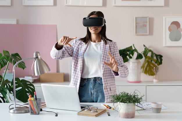 Foto fiduciosa giovane donna in cuffia per realtà virtuale che punta in aria mentre si trova al suo posto di lavoro in ufficio