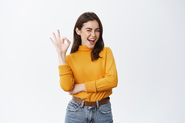 Confident young woman winking to assure you showing okay sign and smiling guarantee good quality say alright or agree like something or recommending white background