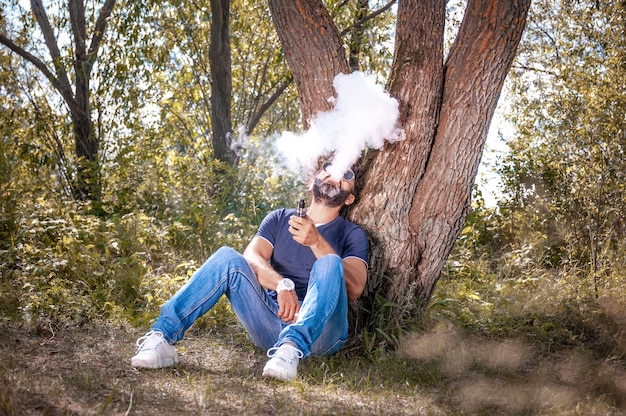 L'uomo fiducioso sta riposando fuori e svapando e scaricando vapore da un dispositivo elettronico per il fumo. t