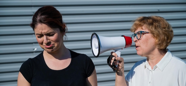 The conflict of generations An old woman yells into a megaphone at her middleaged daughter against gray background Quarrel between an elderly mother and daughter